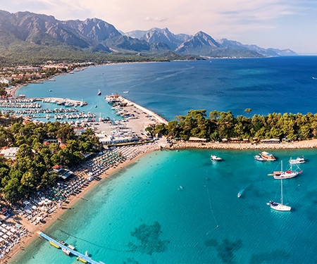 <p>Turkooiskleurige baai met strand en aangemeerde boten, omringd door groen en bergen op de achtergrond, gezien vanuit de lucht.</p>