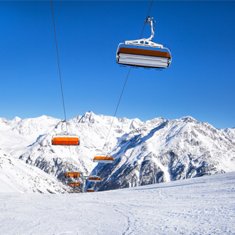 <p><span>Stoeltjeslift met oranje zitjes zweeft boven een besneeuwde skipiste, met hoge bergtoppen op de achtergrond en een strakblauwe lucht.</span></p>