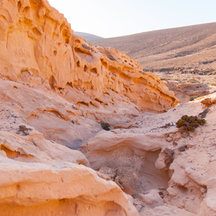 Zandkleurige rotsformatie in een droog woestijnlandschap, met uitgesleten vormen en een smalle doorgang tussen de rotsen.