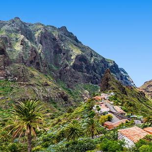 Bergdorp Masca tussen hoge rotswanden, met palmbomen, terracotta daken en kronkelende weg in een groene vallei.