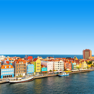 Kleurrijke koloniale huizen langs de waterkant van Willemstad op Curaçao, met de Handelskade en een kalme baai onder een heldere blauwe lucht.