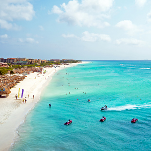 Lang wit zandstrand langs een turquoise zee in Aruba, met zwemmers, waterscooters en strandbedden verspreid langs de kust.