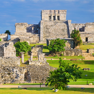 Oude stenen ruïnes van tempels op een groen terrein, met lage muren, trappen en bomen, gezien onder een heldere blauwe lucht.
