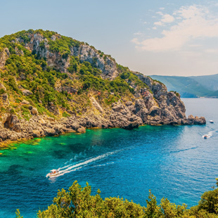 Baai van Paleokastritsa met groen begroeide rotsen, helderblauw zee water en een speedboot die een wit spoor achterlaat.