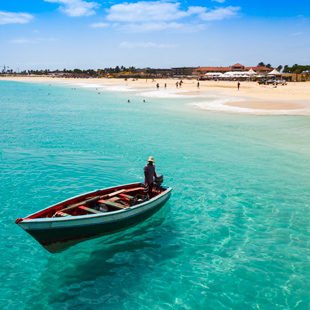 Klein houten bootje met een visser in helder turquoise water, met een wit zandstrand en lage bebouwing op de achtergrond in Kaapverdië.