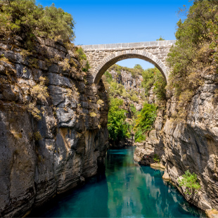Oude stenen boogbrug overspant een smalle kloof met helder groenblauw water, omringd door rotswanden en bomen.