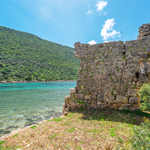Oude stenen ruïnemuur aan een rustige turquoise baai, met groene heuvels op de achtergrond en helder water langs de rotsachtige kust.