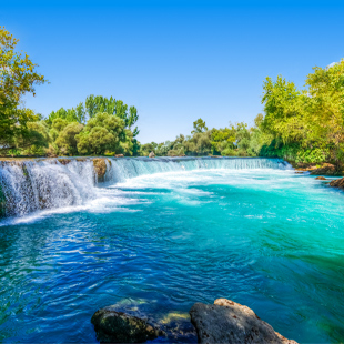 Brede waterval stroomt in een helder turquoise rivier, omringd door groene bomen en rotsen onder een zonnige blauwe lucht.