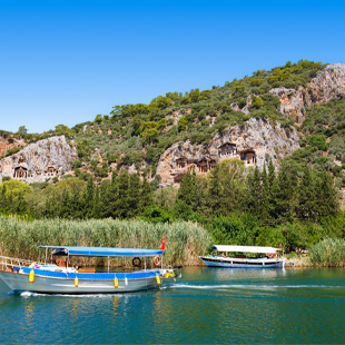 Kleine boten varen over een rustige rivier met riet langs de oever, met rotswanden en oude rotsgraven in het groene landschap.